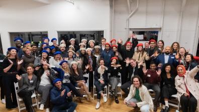 Group photo of attendees at the Columbia Convening on AI Openness and Safety, smiling and waving while wearing blue, red, and white berets, seated and standing in a brightly lit room with large windows.